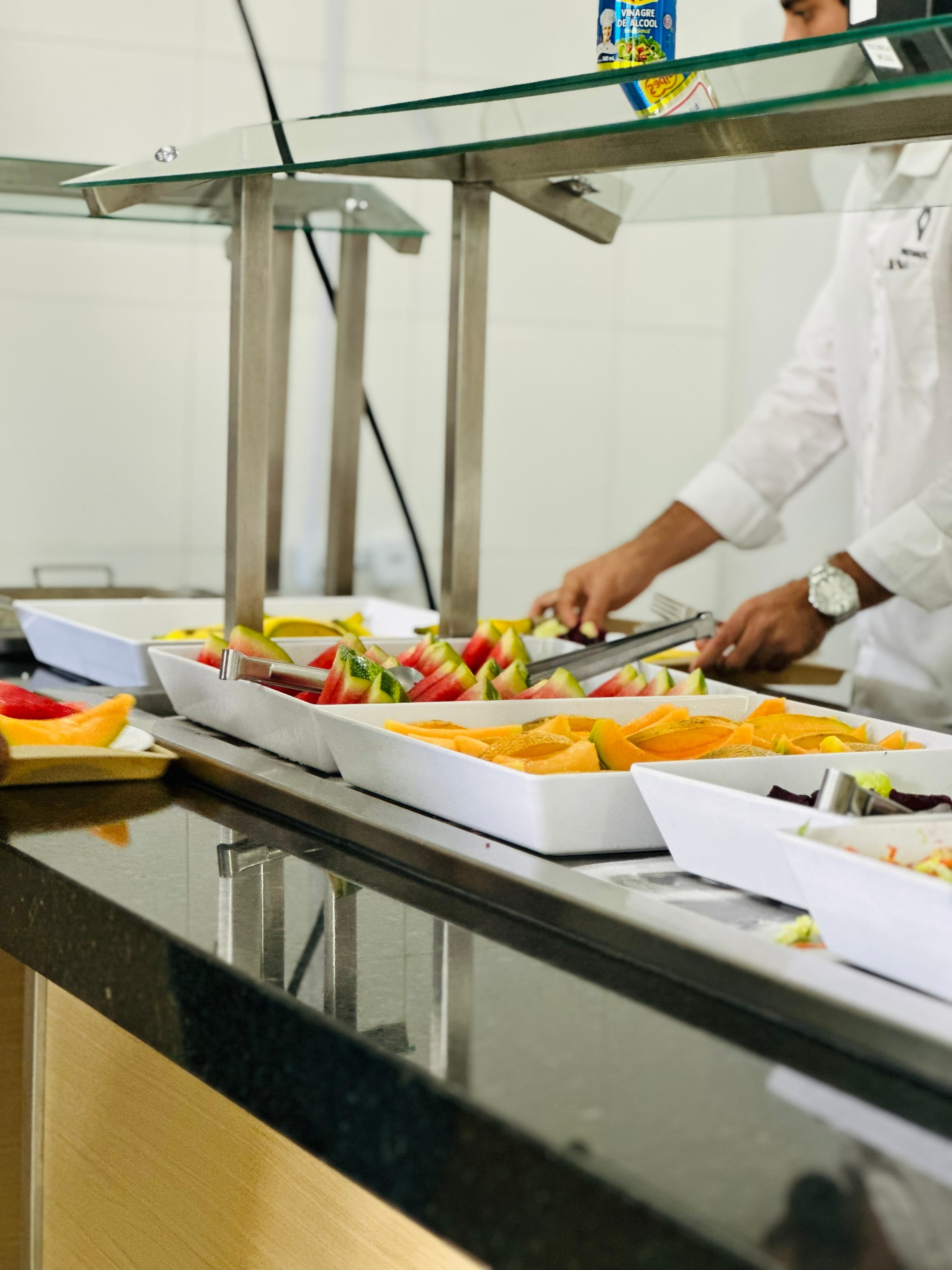 Buffet de frutas com melancia e melão sendo preparados por um cozinheiro em ambiente de cozinha profissional.