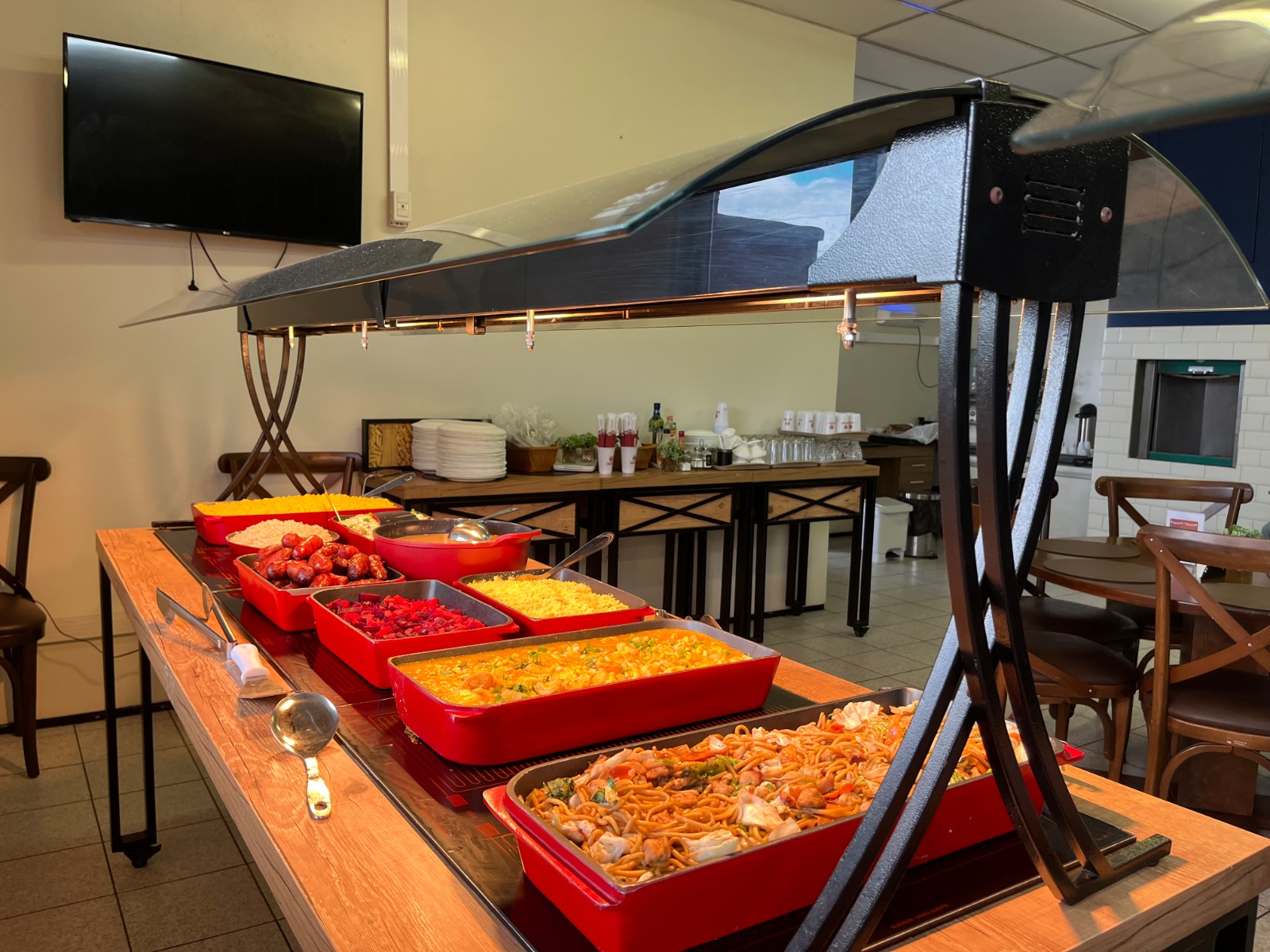 Visão ampla de um buffet self-service com várias opções de pratos quentes em recipientes vermelhos sobre balcão aquecido.