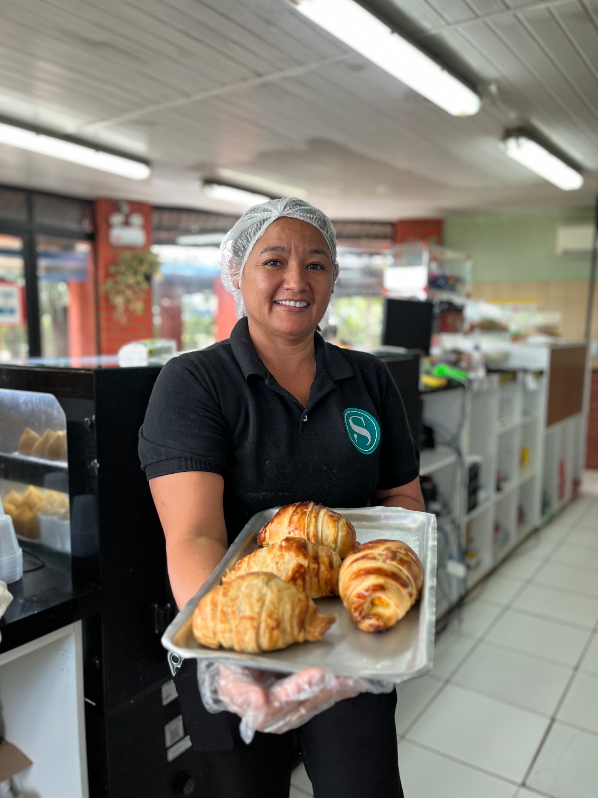 Funcionária de padaria sorrindo, usando touca e luvas, segurando uma bandeja com pães frescos.